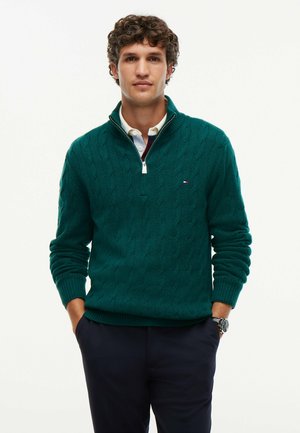 Homme aux cheveux bouclés portant un pull vert en maille torsadée à fermeture éclair sur le quart, par-dessus une chemise blanche à col, les mains dans les poches d’un pantalon bleu marine.