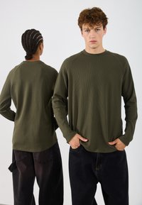 Olīvu zaļa garām piedurknēm krekls ar strukturētu ribu dizainu, apaļu kakla izgriezumu un raglāna piedurknēm, kombinēts ar tumšiem, brīvi pieguļošiem biksēm.