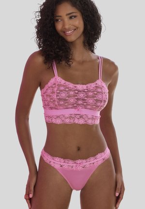 Frau mit lockigem Haar, die lächelt und ein passendes pinkfarbenes Blumen-Spitzen-Bralette und Tanga trägt, vor einem einfachen grauen Hintergrund.