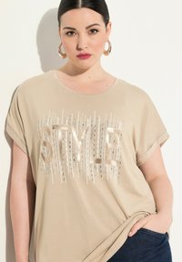 Béžové tričko s krátkým rukávem s metalickým designem textu "STYLE" a dekorativními akcenty. Hladká tkanina, uvolněný střih, jemná textura.