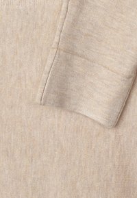 Tissu beige avec une texture douce, présentant un poignet côtelé. La surface montre un motif subtil et irrégulier, typique des matériaux tricotés.
