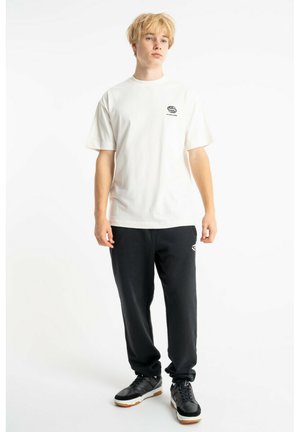 Młody mężczyzna z blond włosami, ubrany w białą koszulkę z nadrukiem, czarne spodnie dresowe i czarne trampki, stojący na białym tle.