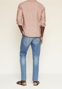 Camisa a rayas de manga larga, marrón rojizo, con los puños remangados, combinada con unos vaqueros rectos de color azul claro y sandalias marrones.