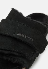 Black suede sandals with two adjustable straps, featuring a soft, fuzzy interior. The brand name "Birkenstock" is visible on the strap.