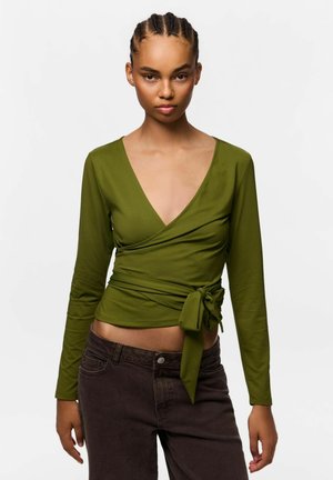 Femme aux cheveux tressés portant un haut cache-cœur vert olive noué à la taille et un pantalon marron foncé, debout devant un fond blanc uni.
