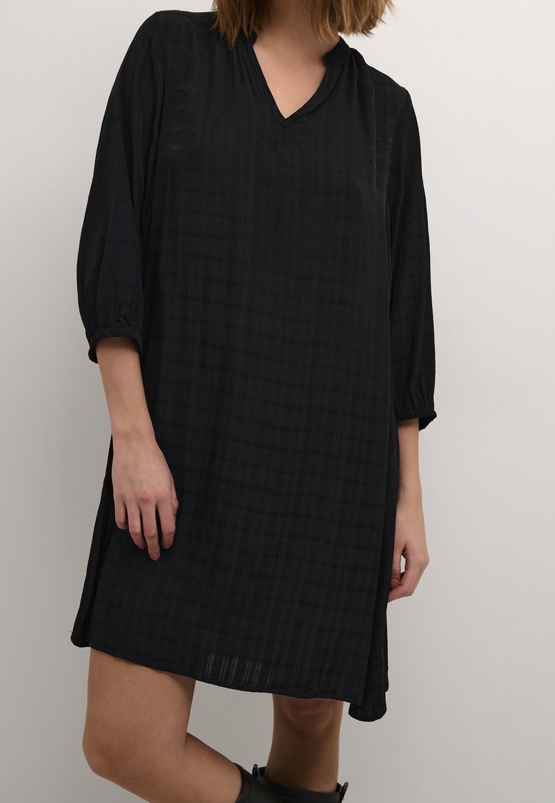 Femme portant une robe noire ample à manches trois-quarts, avec une texture discrètement à carreaux, debout devant un fond clair et uni.