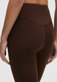 Bruine high-waisted leggings gemaakt van glad, rekbaar materiaal met platte naden en een klein logo-detail op de achterkant.