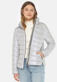 Hellgraue Pufferjacke mit glänzender Oberfläche, horizontalem Steppmuster, hohem Kragen, Reißverschluss und seitlichen Taschen. Über einem weißen Oberteil getragen.