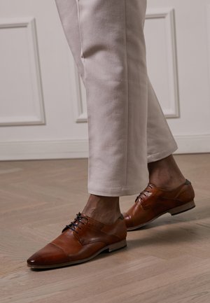 Brown leather dress shoes with a pointed toe, textured accents, and black laces, paired with light cream-colored trousers.
