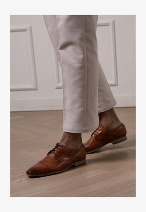 Brown leather dress shoes with a pointed toe, textured accents, and black laces, paired with light cream-colored trousers.