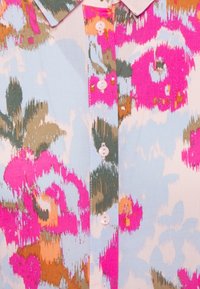 Bloes met bloemenpatroon in roze, groen en oranje op een lichtblauwe achtergrond. Heeft een kraag en kleine, ronde, roze knopen aan de voorkant.