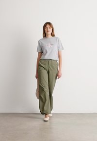 Mujer caminando en interiores, vestida con una camiseta gris de Calvin Klein, pantalones de pierna ancha en verde oliva, zapatos blancos y sosteniendo un bolso redondo beige.