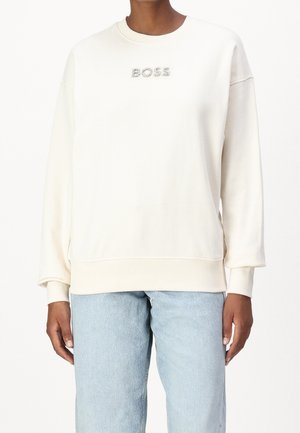 Osoba ubrana w białą bluzę z napisem „BOSS” wykonanym z dżetów na piersi oraz jasnoniebieskie dżinsy, stojąca na białym tle.