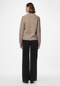 Gestrickter Pullover in hellem Beige mit strukturiertem Muster, Rundhalsausschnitt und Zopfmuster-Ärmeln. Kombiniert mit hoch taillierter schwarzer Hose.