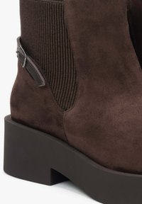 Bruine suède enkelboot met ribbelstof accenten, een platte gelamineerde zool en een zilverkleurige gespdetail aan de zijkant.