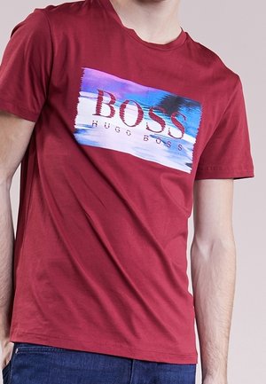 Mann trägt rotes Hugo Boss T-Shirt mit blauem, weißem und violettem Farbstrich-Design und Logo auf der Brust.