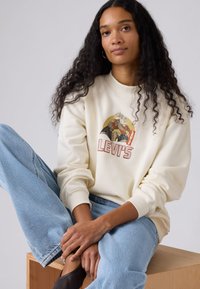 Felpa color crema con stampa grafica e logo Levi's rosso, abbinata a jeans azzurri, che mostrano una vestibilità comoda e polsini a coste.