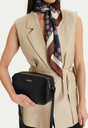 Kobieta w beżowej marynarce bez rękawów i wzorzystym szaliku w kolorach granatowym, białym i brązowym, trzymająca czarną teksturowaną skórzaną torbę na ramię z suwakiem.