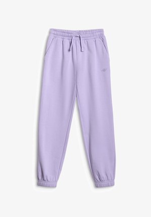 Jong meisje met lang bruin haar, een witte haarband, een crèmekleurige sweater met ritssluiting, lavendelkleurige joggingbroek en witte sportschoenen, staand met handen in de zakken.