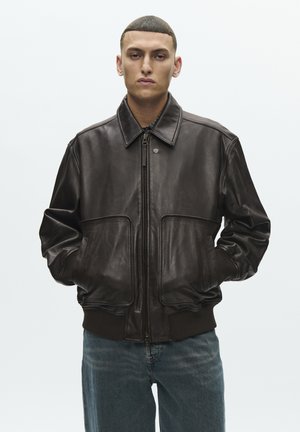 Férfi áll zsebében tartott kézzel, sötétbarna bőr cipzáras dzsekit és kék farmert viselve, világos háttér előtt.