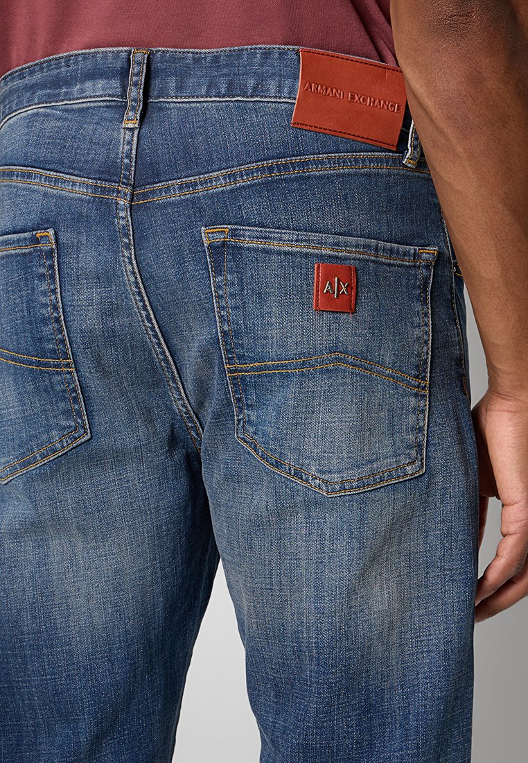 Primo piano di una persona che indossa jeans blu Armani Exchange con toppe rosse sui taschini posteriori e sulla cintura.