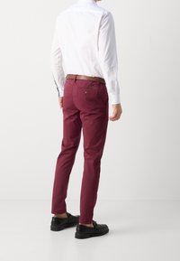 Chinos bordeaux con una vestibilità slim, dotati di una tasca posteriore e una cintura in pelle marrone, abbinati a scarpe nere e una camicia bianca a maniche lunghe.