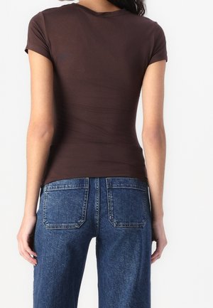 Woman wearing a fitted brown short-sleeve top and high-waisted blue denim jeans, shown from the back against a plain background.