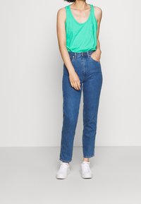 Canotta verde con tasca, abbinata a jeans blu a vita alta con taglio dritto e sneakers bianche. Design semplice.