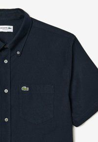 Camicia button-up a maniche corte di colore blu navy in cotone, con taschino sul lato sinistro del petto e un piccolo logo di coccodrillo ricamato in verde sulla tasca.