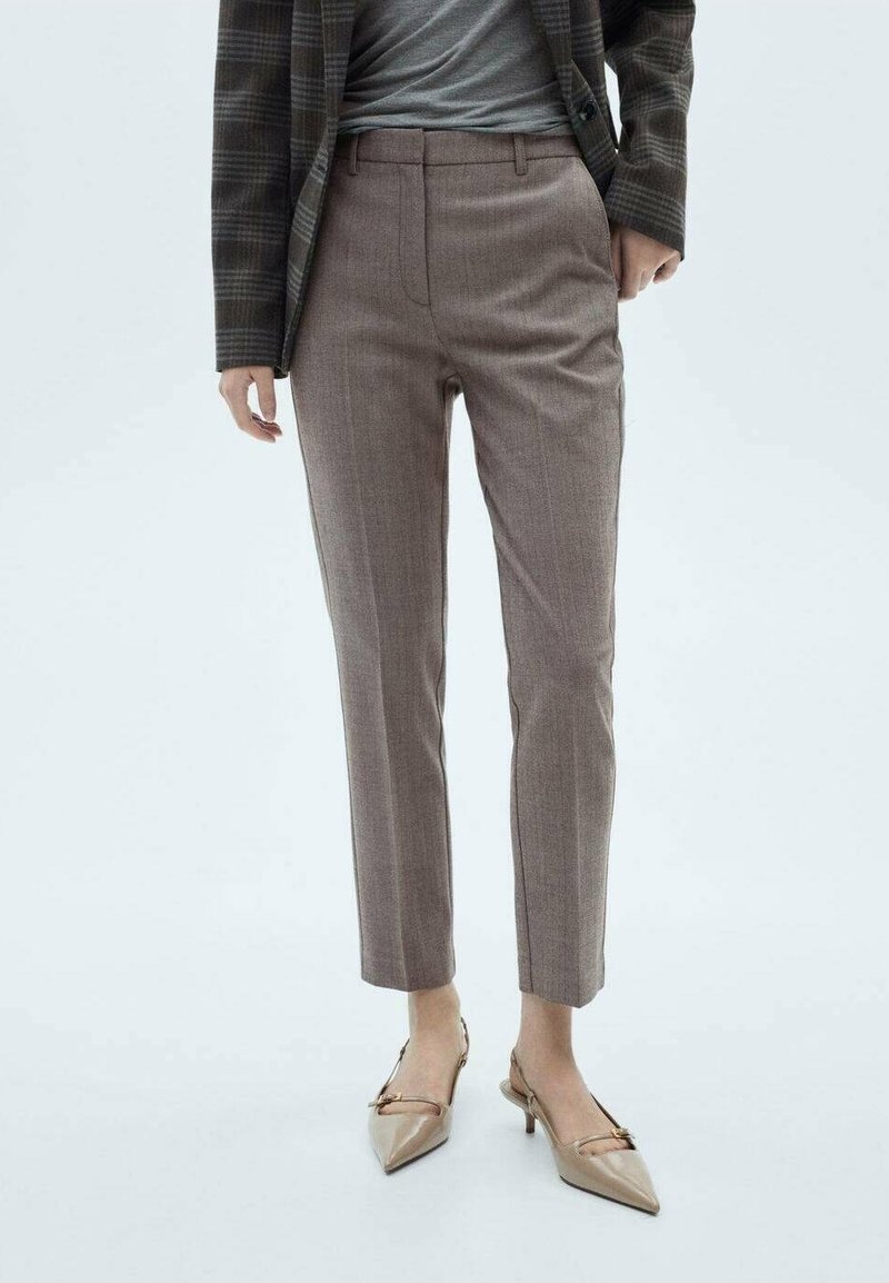 Personne portant un blazer à carreaux gris, un haut gris, un pantalon marron tailleur et des chaussures beiges pointues à talon bas, debout sur un fond blanc.