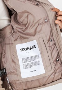 Beige pufferjacka med insidan täckt av "SIXTH JUNE"-tryck. Har en vit etikett med varumärkesinformation och två tryckknappar.
