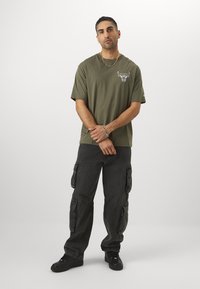 Olivengrøn t-shirt med logo på brystet, parret med sorte cargo-bukser med flere lommer og sorte sneakers. Enkle smykkedetaljer.