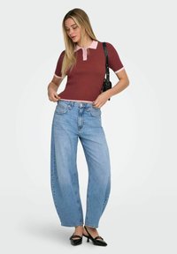 Jeune femme portant un polo côtelé bordeaux, un jean large bleu, des chaussures à talons noires, et portant un sac à épaule noir.