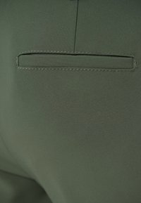 Țesătură verde închis, cu o textură netedă; prezintă un buzunar din spate cu detalii de cusătură dreaptă și curată, fără hardware vizibil.