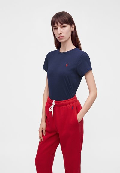 Γυναίκα που φοράει μπλε σκούρο T-shirt και κόκκινο παντελόνι φόρμας με λευκό κορδόνι, στέκεται με το ένα χέρι στην τσέπη μπροστά σε απλό ανοιχτό φόντο.