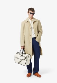 Trench-coat beige porté sur un polo blanc, jean bleu foncé, baskets orange, tenant un sac de sport blanc avec des accents noirs et le texte "LACOSTE".