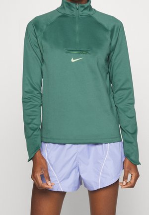 Personne portant un pull Nike vert à manches longues avec une poche zippée à l'avant et un short de course violet clair.
