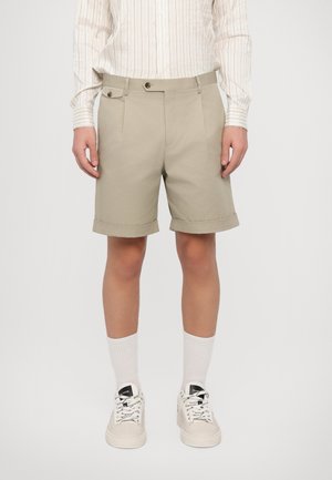 Beige Shorts mit umgeschlagenem Saum, ausgestattet mit frontalen Falten und Knopfdetails, kombiniert mit weißen Socken und hellbeigen Sneakers.