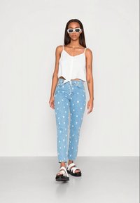 Top branco cropped com botões e laço na cintura, jeans de ganga azul claro com bolinhas brancas e sandálias pretas com tiras brancas.
