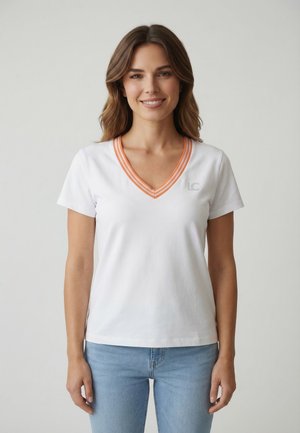 Mujer con camiseta blanca de cuello en V con borde naranja y vaqueros azul claro, sonriendo, de pie frente a un fondo claro y liso.