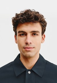 Czarna koszula z kołnierzykiem o gładkiej fakturze, z zapięciem na guziki. Zbliżenie na młodego mężczyznę z kręconymi włosami i neutralnym tłem.