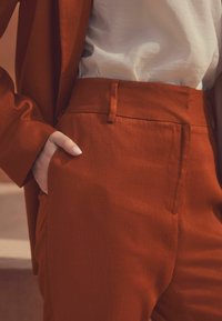 Pantalons en lin couleur rouille avec une texture lisse, dotés d'une taille ajustée, de poches latérales et d'une veste assortie. Haut blanc en dessous.