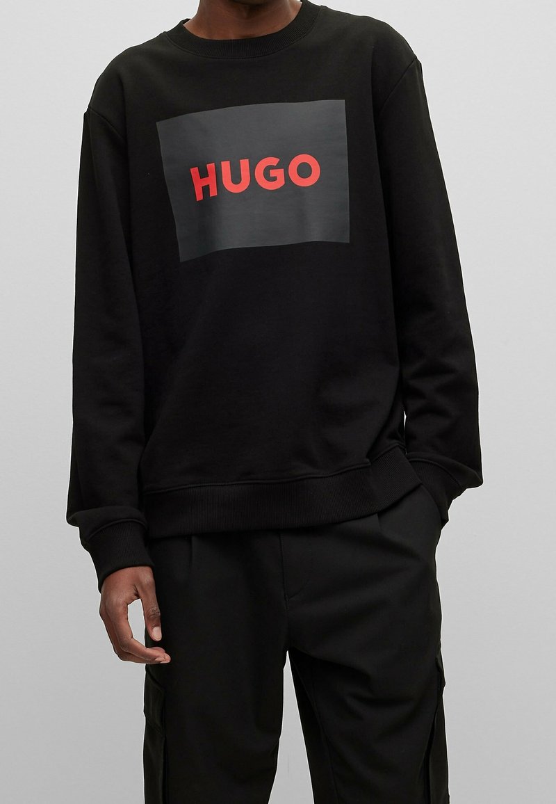 Czarna bluza z prostokątnym nadrukiem z napisem "HUGO" w czerwonym kolorze. Miękka tkanina, prążkowany dekolt i mankiety, luźny krój.