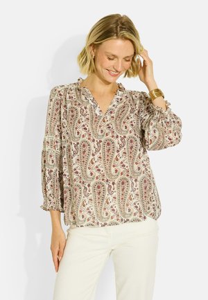 Mujer sonriendo y tocándose el pelo, lleva una blusa crema con estampado paisley y mangas tres cuartos, y pantalones blancos.