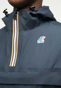 Κοντινό πλάνο σκούρου μπλε αδιάβροχου μπουφάν με φερμουάρ σε ρίγα και λογότυπο K-Way στο στήθος, με ρυθμιζόμενο κουκούλα με κορδόνια.