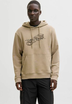 Mann trägt einen beigen Kapuzenpullover mit schwarzem, geschichtetem Textgrafik und schwarze Cargo-Hosen, steht vor einem schlichten hellen Hintergrund.