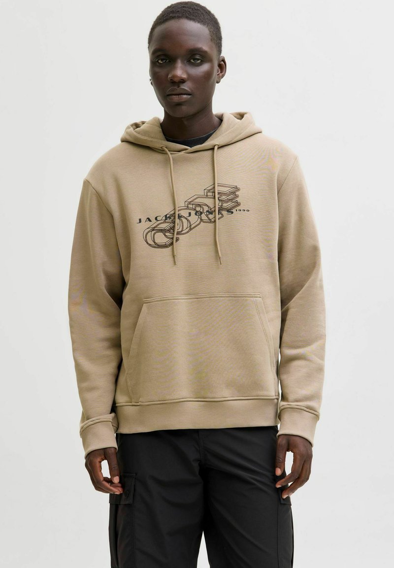 Mężczyzna w beżowej bluzie z kapturem z czarnym nadrukiem warstwowym i czarnych spodniach cargo stoi na tle jasnego jednolitego tła.