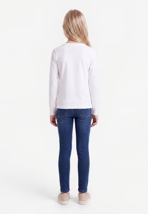 Bambino con lunghi capelli biondi che indossa una maglietta bianca a maniche lunghe, jeans blu e sneaker rosa chiaro, in piedi di spalle su sfondo bianco.