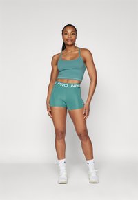 Grønn sportsbh med justerbare stropper, kombinert med matchende høyde shorts med hvit "PRO" midje. Hvite joggesko og sokker.
