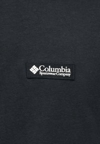 Κονκάρδα με το λογότυπο της Columbia Sportswear Company σε λευκό χρώμα πάνω σε μαύρο υφασμάτινο φόντο.
